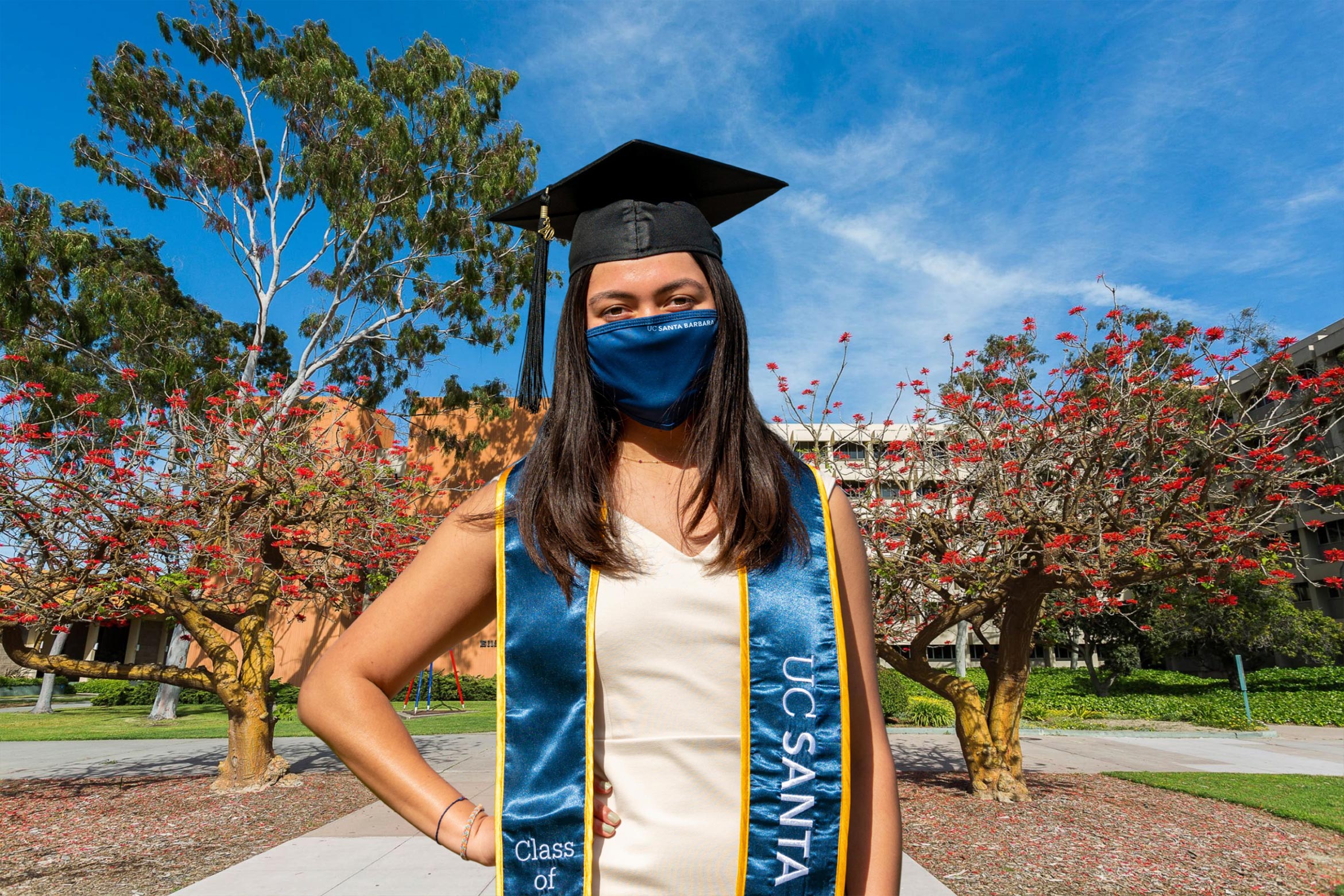 A masked student with graduation regalia poses against a backdrop of coral trees on the UC Santa Barbara campus.