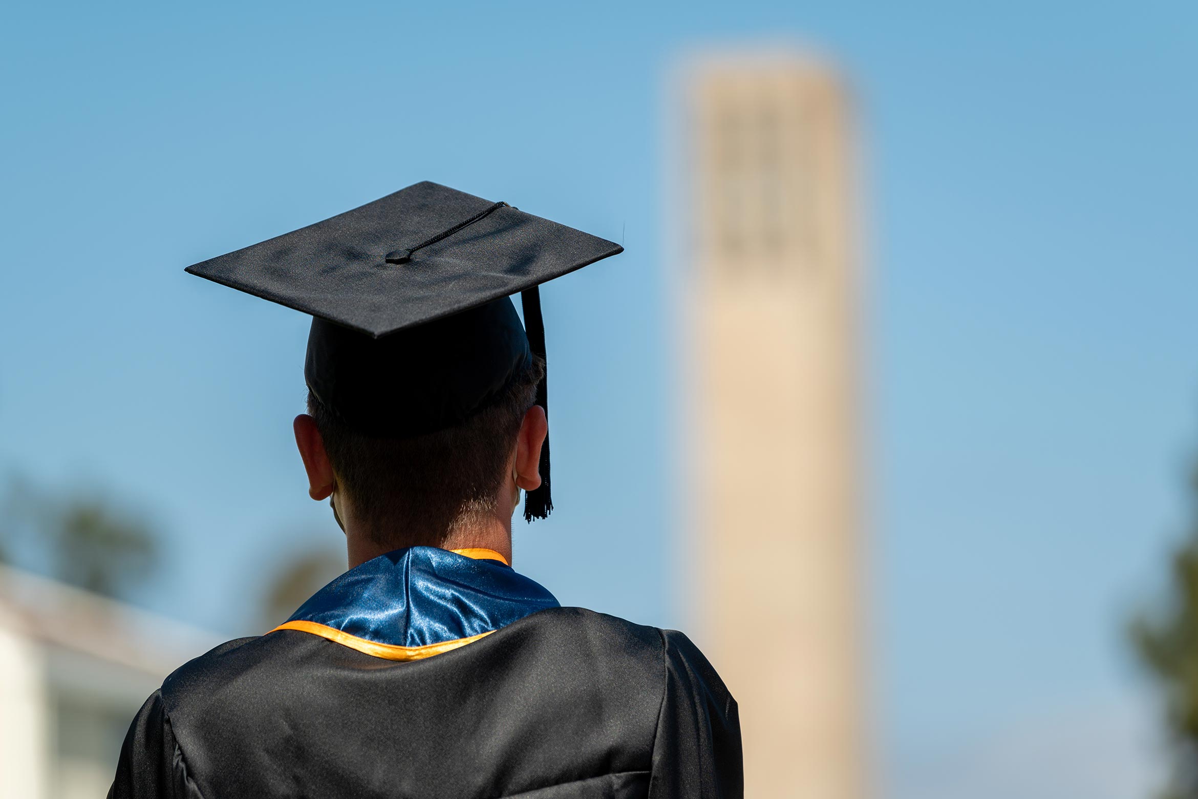 A UC Santa Barbara graduate wearing a cap, gown, and stole stands on campus looking at Storke Tower in the distance.